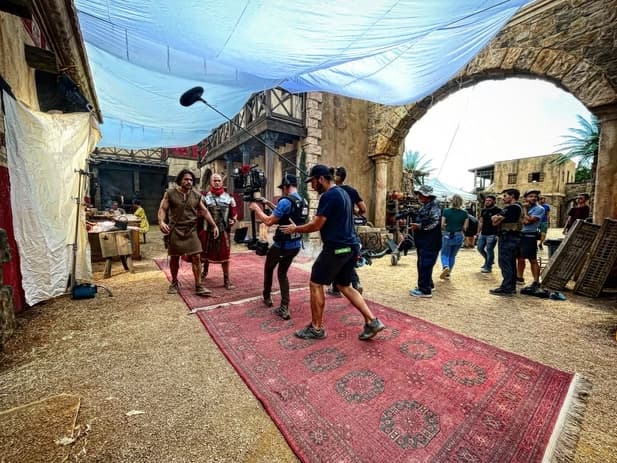 A camera team filming performers on a large exterior set